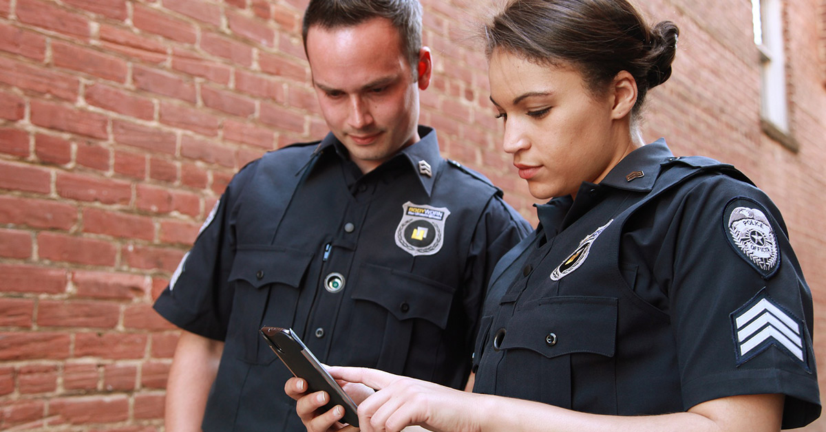 A female uniformed police officer holds a smart phone pointing to the screen while a male uniformed police officer looks on with interest.