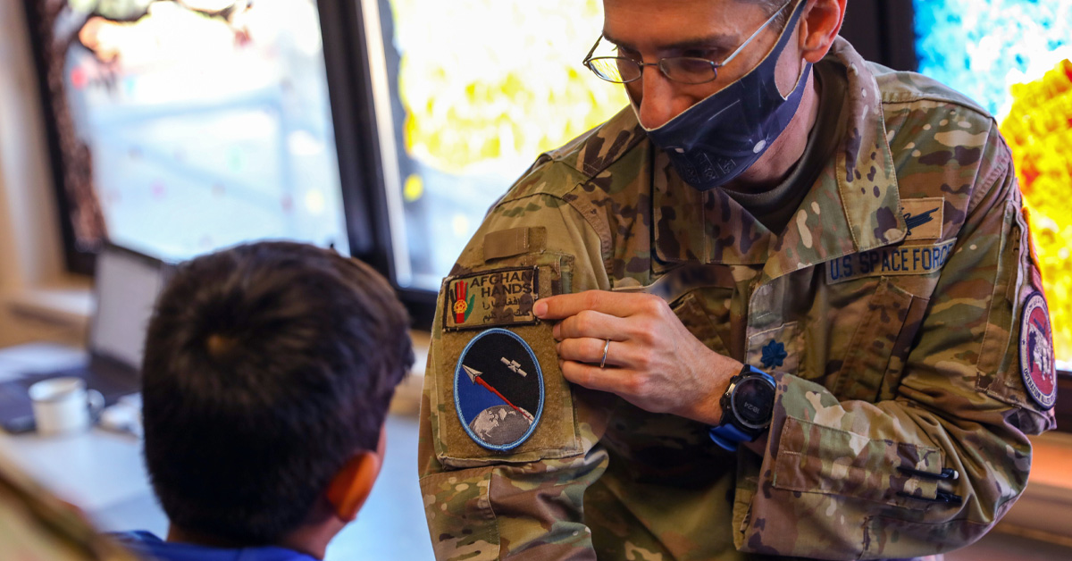 A male soldier wearing army fatigues and a face mask showing his space force patch on his arm to a child.