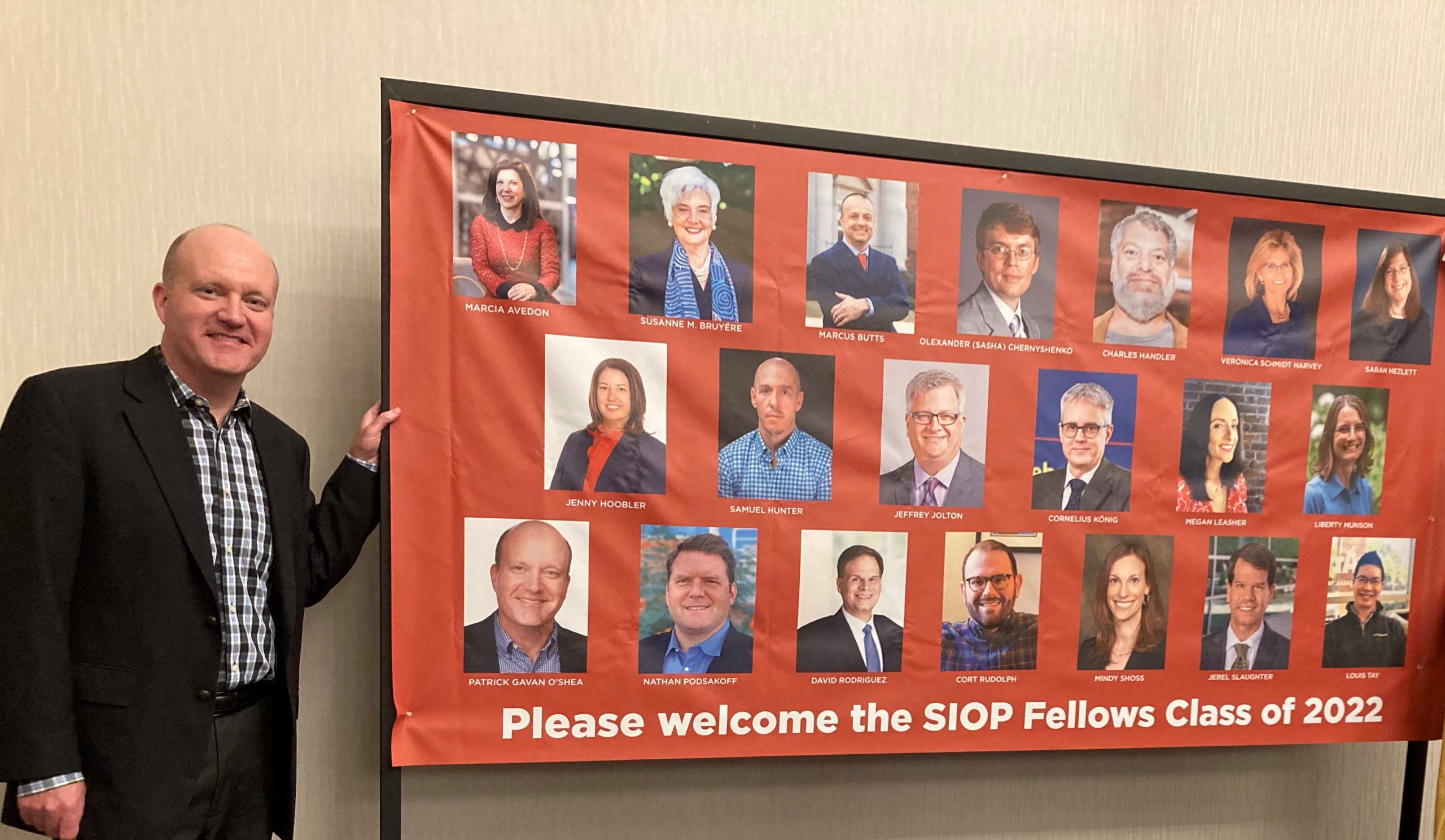 Gavan O'Shea smiling and standing next to a board with pictures of all Fellow board members