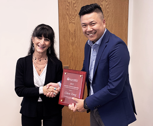 Suzanne Tsacoumis and Chen Tang shaking hands, Chen Tang holding and displaying the award